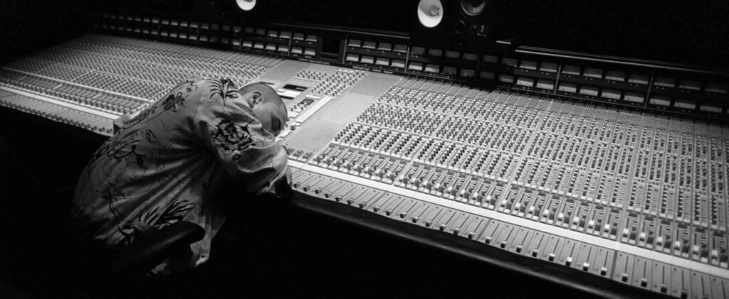 A young man sleeping against a large sound mixing board 